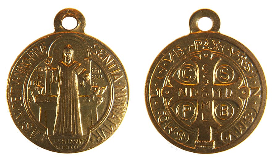 守護靈魂的古老盾牌：聖本篤勳章（Saint Benedict Medal）這是一張展示兩枚金色圓形聖本篤勳章正反兩面的特寫照片。左側為勳章正面，刻有一名站立的聖徒（聖本篤），他右手握著十字架，左手持書，背景有祭壇與拉丁銘文。右側為勳章反面，中心是一個大十字架，十字架與圓周邊緣刻有多組拉丁文首字母縮寫（如 C S S M L, N D S M D），頂端刻有「PAX」（和平）字樣。勳章頂部皆有圓孔掛環，呈現金屬光澤與細緻的浮雕工藝。