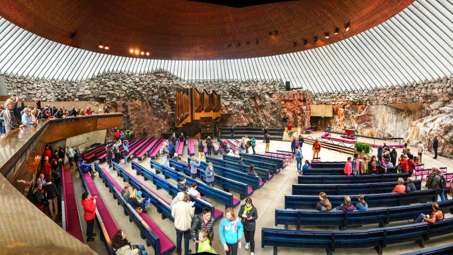芬蘭赫爾辛基岩石教堂（Temppeliaukio Church）內部景觀，呈現粗獷的天然花崗岩牆面、巨大的圓形銅製屋頂與環繞的透光窗櫺，藍色長椅環繞著中央祭壇空間。